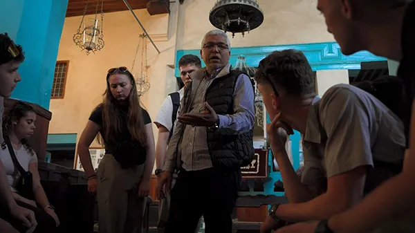 A person wearing glasses gestures while speaking to a group gathered inside a room with turquoise walls and hanging lanterns.