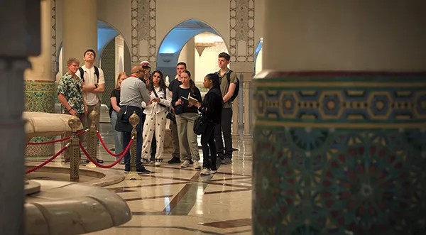 A person gestures while speaking to a group inside a hall with arched doorways, tiled columns, and patterned floors.