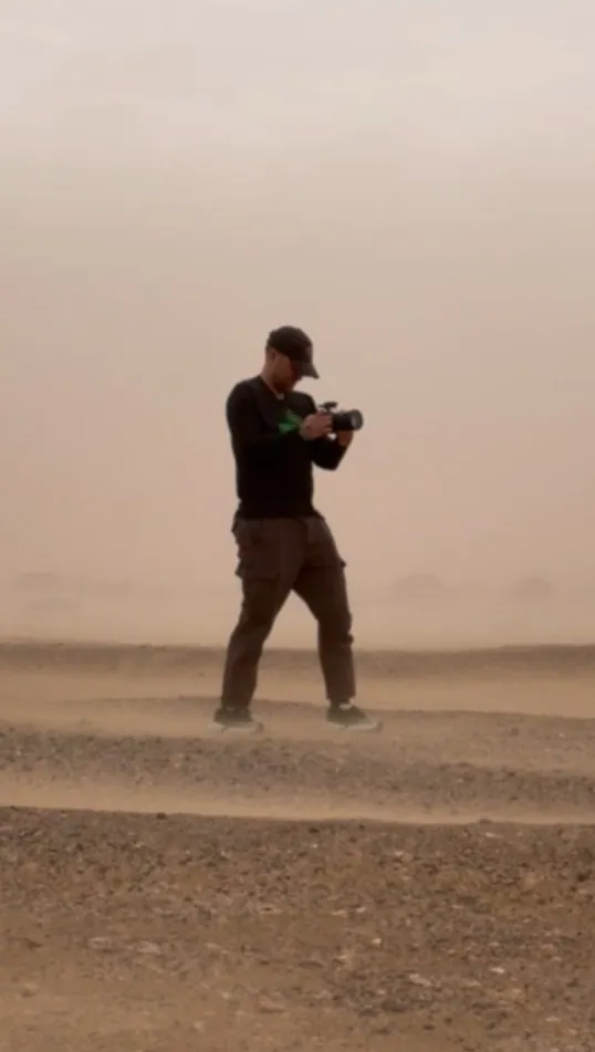 A person wearing a cap holds a camera while walking across a dusty, rocky landscape.