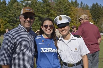 Three people stand together on a field, including a cadet in uniform, smiling for a photo.