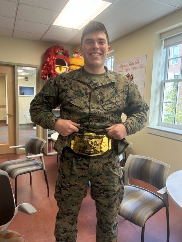 A Norwich University cadet proudly displays a gold “Language Slayer” championship belt, celebrating academic excellence.