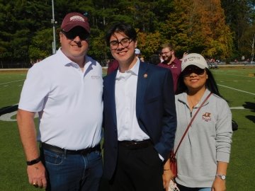 Three people stand together on a field wearing Norwich University apparel, smiling for a photo.