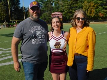 Cheerleader in uniform stands on a football field with two people wearing Norwich University apparel.