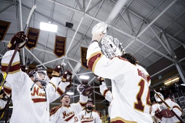 Two hockey players in maroon and gold uniforms, one holding up a trophy, celebrating a victory on the ice with teammates in the background under banners.