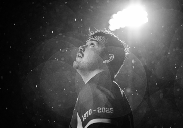 Close-up of an athlete in a jersey marked '1970 - 2025' looking upward with visible water droplets in the air, illuminated by a bright light overhead.