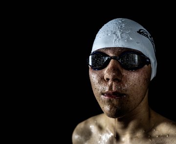 A swimmer wearing a Speedo cap and goggles, with water droplets on their face, against a dark background.