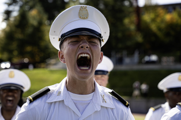 A cadet in a white uniform with a visor cap is shouting orders, with other cadets visible in the background.