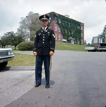 corporal cadet in uniform standing in parking lot before several cars and ivy covered building