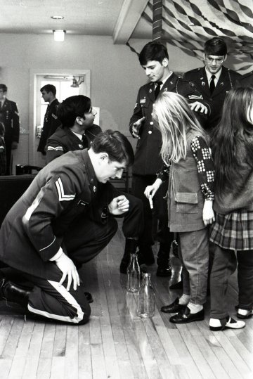 cadet in uniform kneels before two children, with two other cadets in uniform beside them