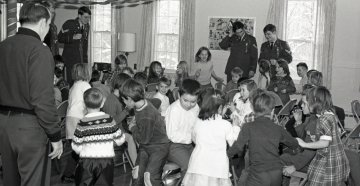 a crowd of children are watch by a man (seen from his back) and three Norwich cadets in uniform