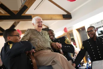 A joyful senior in a glittery top laughing heartily while being playfully carried by three smiling people in military uniforms in a festive room with balloons and wooden beams.
