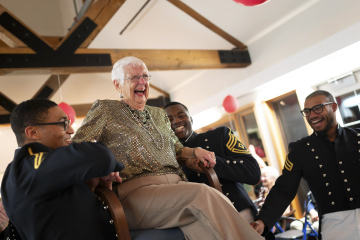 A joyful senior in a glittery top laughing heartily while being playfully carried by three smiling people in military uniforms in a festive room with balloons and wooden beams.
