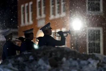 Three members of the Norwich University Corps of Cadets playing trumpets outdoors at night with snow falling, illuminated by a street lamp in front of a brick building.