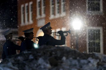 Three members of the Norwich University Corps of Cadets playing trumpets outdoors at night with snow falling, illuminated by a street lamp in front of a brick building.