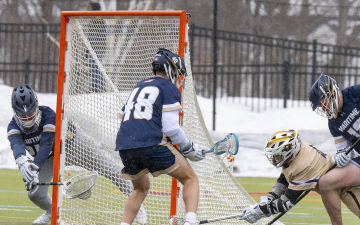 Lacrosse players engaged in a game, with one player shooting on goal while a goalie attempts a save and another defends.