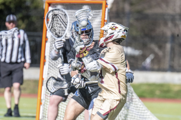 Two lacrosse players are competing near the goal during a game, with one player cradling the ball and the other attempting to block.