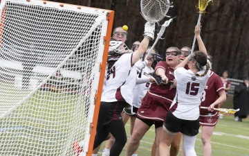 Lacrosse players from two teams in action near the goal, attempting to catch or block a yellow ball.