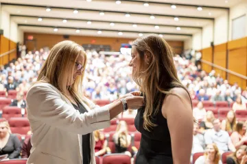 Two individuals on stage in a conversation, with one placing a pin on the other, in front of an audience in a lecture hall.