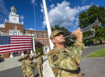 A member of the Norwich University Corps of Cadets plays a trumpet in front of a building with a large American flag. Another uniformed personnel is partially visible in the background.