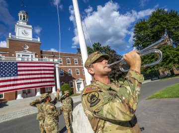 A member of the Norwich University Corps of Cadets plays a trumpet in front of a building with a large American flag. Another uniformed personnel is partially visible in the background.