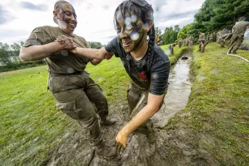 Two soldiers participating in a muddy training exercise, one assisting the other in navigating through the mud, both wearing camouflage uniforms with a red poppy symbol.