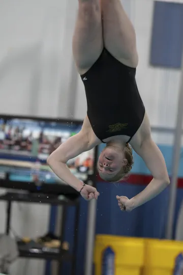 A diver from Norwich University performs an inverted flip during a routine, captured mid-air against a backdrop of spectators and pool equipment.