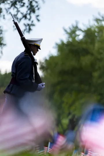 A member of the Norwich University Corps of Cadets in dress uniform walking solemnly past a field of American flags.
