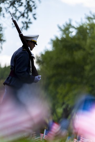 A member of the Norwich University Corps of Cadets in dress uniform walking solemnly past a field of American flags.