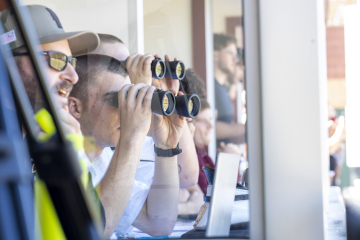 Spectators using binoculars to watch an event from behind a window.