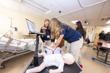 Three healthcare trainees collaborate around a medical training mannequin in a hospital simulation room, engaging with computer equipment to monitor the simulation.
