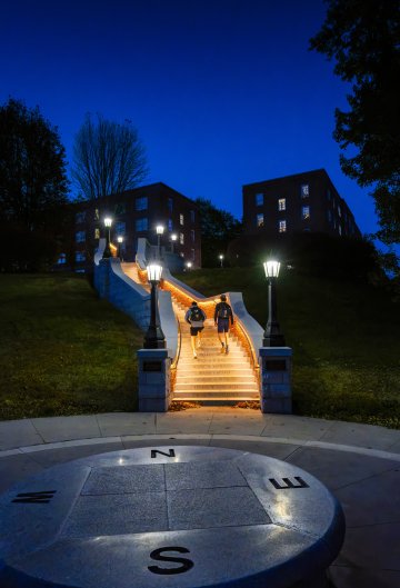 Three individuals walking up a lit staircase at twilight, with a compass rose in the foreground and buildings in the background.