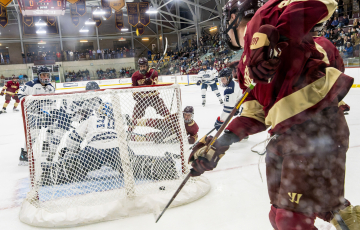 Hockey player from team in maroon and gold uniform attempting a goal against a goalie from team in white and blue, during a fast-paced game.