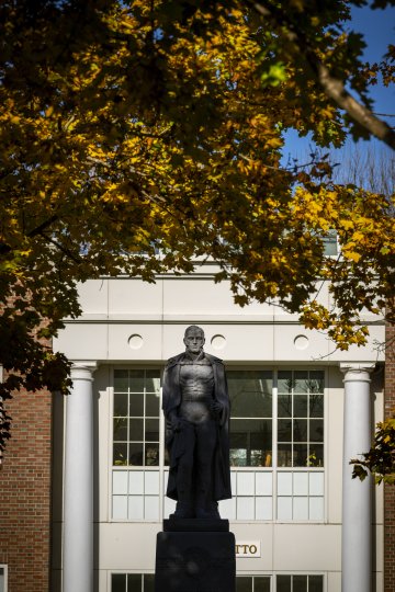 A statue of Alden Partridge stands in the foreground framed by autumn-colored leaves, with a classic building featuring white columns in the background.