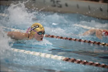 Swimmer wearing a yellow "Norwich" cap competes in a breaststroke race in an indoor pool.