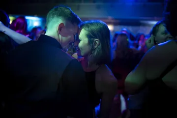 Two people sharing an intimate moment on a dance floor with dim blue lighting and a crowd around them.