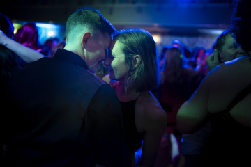Two people sharing an intimate moment on a dance floor with dim blue lighting and a crowd around them.