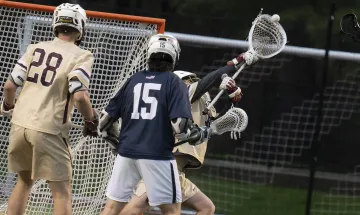 A lacrosse game in progress with three players in action; one player in a white and gold uniform marked "28" and another in dark blue marked "15" are observing a third player in a white jersey, also marked "15," making a save as a lacrosse ball flies towards the goal.