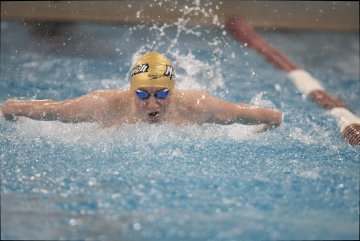 Swimmer in action at a pool wearing a cap with 'Norwich' logo and blue goggles, competing in a butterfly stroke race.