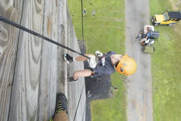 A person wearing an orange helmet and gloves is rappelling down a tall wooden structure, with several onlookers and a vehicle visible on the ground below.