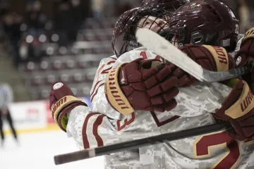 Two Norwich University ice hockey players in maroon and gold uniforms embracing joyfully on the rink.