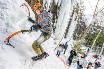 Two individuals practice ice climbing on a frozen waterfall with others observing and assisting from below. One climber is actively ascending with ice axes, wearing a helmet and harness. The scenery includes snow-covered trees and rocks.