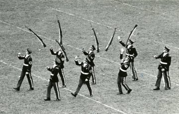 eight men in uniform marching forward throwing rifles above them