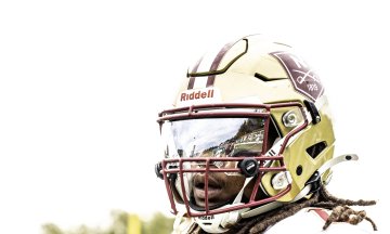 Close-up of a person wearing a Norwich University football helmet with dreadlocks visible, set against a lightly clouded sky.