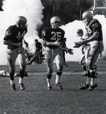 three football players run up the field celebrating together, smoke from a cannon rises in background