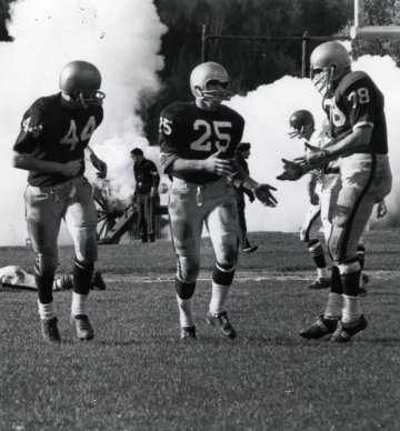 three football players run up the field celebrating together, smoke from a cannon rises in background
