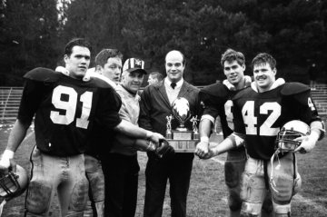 group of four football players in uniform and pads, carrying helmets surround two men, one holding a trophy