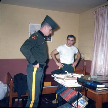 cadet in uniform standing in front of desk with man in white t-shirt in back