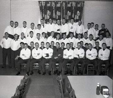 a large group of young men in white shirts sit or stand in three rows, directly in front of a wall and closed curtain