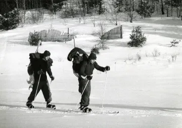 two skiers with backpacks and gear seen crossing meadow