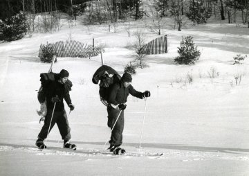 two skiers with backpacks and gear seen crossing meadow